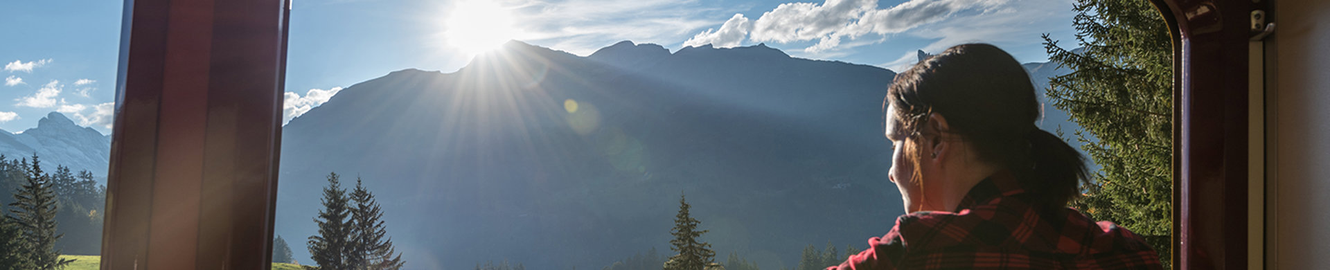 Ung kvinna i röd tröja blickar ut genom tågfönstret mot ett soligt alplandskap.